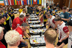 A table hockey tournament hall with rows of tables and national flags overhead